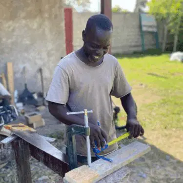 A Haitian carpenter wearing a tan t-shirt smiles as he measures a piece of wood clamped to a workbench outdoors in rural Haiti.