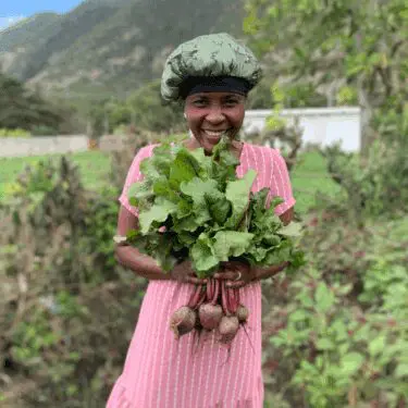 A smiling Haitian woman wearing a pink striped dress and a green patterned chef's cap stands outdoors, holding a freshly harvested bunch of beets with large, vibrant green leaves and reddish-purple roots. A lush green mountain landscape is visible in the background.