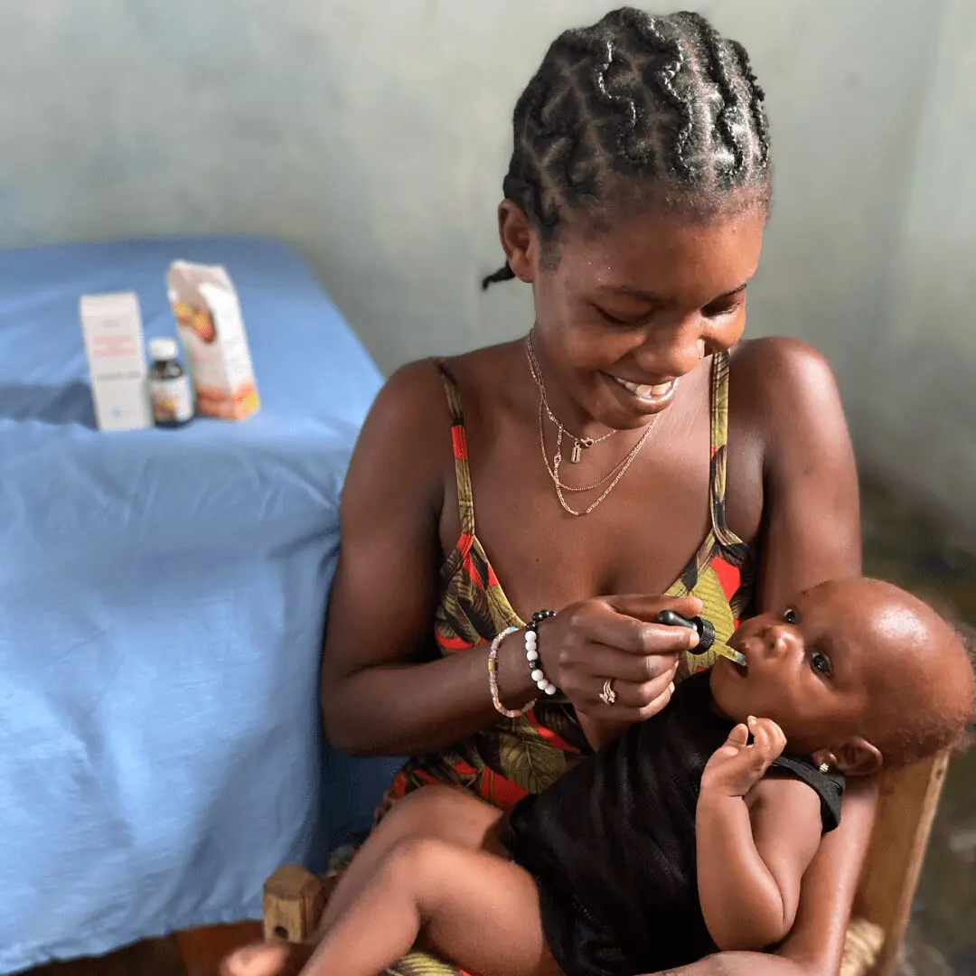 A smiling Haitian woman with braided hair, wearing a patterned tank top, sits and gives a liquid medicine drop to an infant baby she is holding. The baby, dressed in a black outfit, looks up while receiving the medicine. In the blurred background, there's a cot with a blue cover and two medicine bottles on a surface.