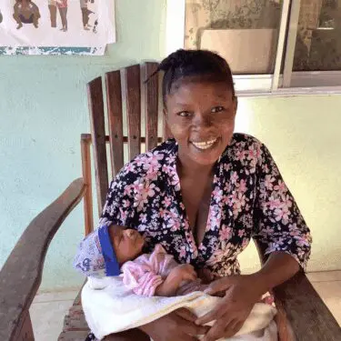 A Haitian woman wearing a black floral robe smiles directly at the camera while sitting in a wooden rocking chair and cradling a newborn baby wrapped in a white blanket and wearing a pink outfit and blue cap. The background is a pale mint green wall with a window and a poster visible on the upper left.