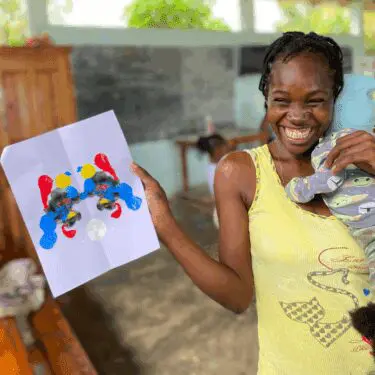 A jubilant Haitian woman wearing a yellow tank top holds a smiling baby wrapped in a gray onesie while proudly displaying a piece of paper with an abstract paint splat design in red, yellow, blue, and black. She is outdoors or under a covered area, with a blurred background showing wooden furniture and a dark wall.