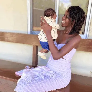 A smiling Haitian woman wearing a light purple strapless dress sits on a wooden bench, holding and gazing lovingly at an infant baby dressed in a cream top with small heart prints. The woman has long, dark twists, and the background is a simple beige wall with a window.
