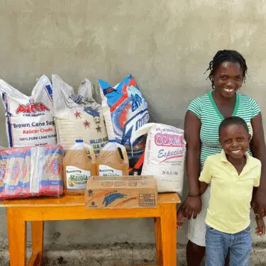 A smiling Haitian woman wearing a green and white striped shirt stands next to a smiling young boy in a yellow polo shirt and jeans, holding hands on either side of a wooden table piled with bags of food aid. The supplies include large sacks of brown cane sugar, rice, flour, cooking oil, and packaged crackers, set against a plain concrete wall.