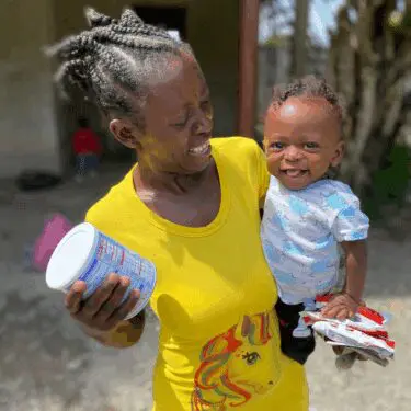 A Haitian woman wearing a bright yellow shirt with a rainbow unicorn graphic holds a happy, smiling baby boy. The baby wears a light blue and white patterned shirt. The woman is looking at the baby, who is looking directly at the camera. In her hand, she holds a white and blue container of formula and nutritional food. They are standing outdoors in a sunny area.