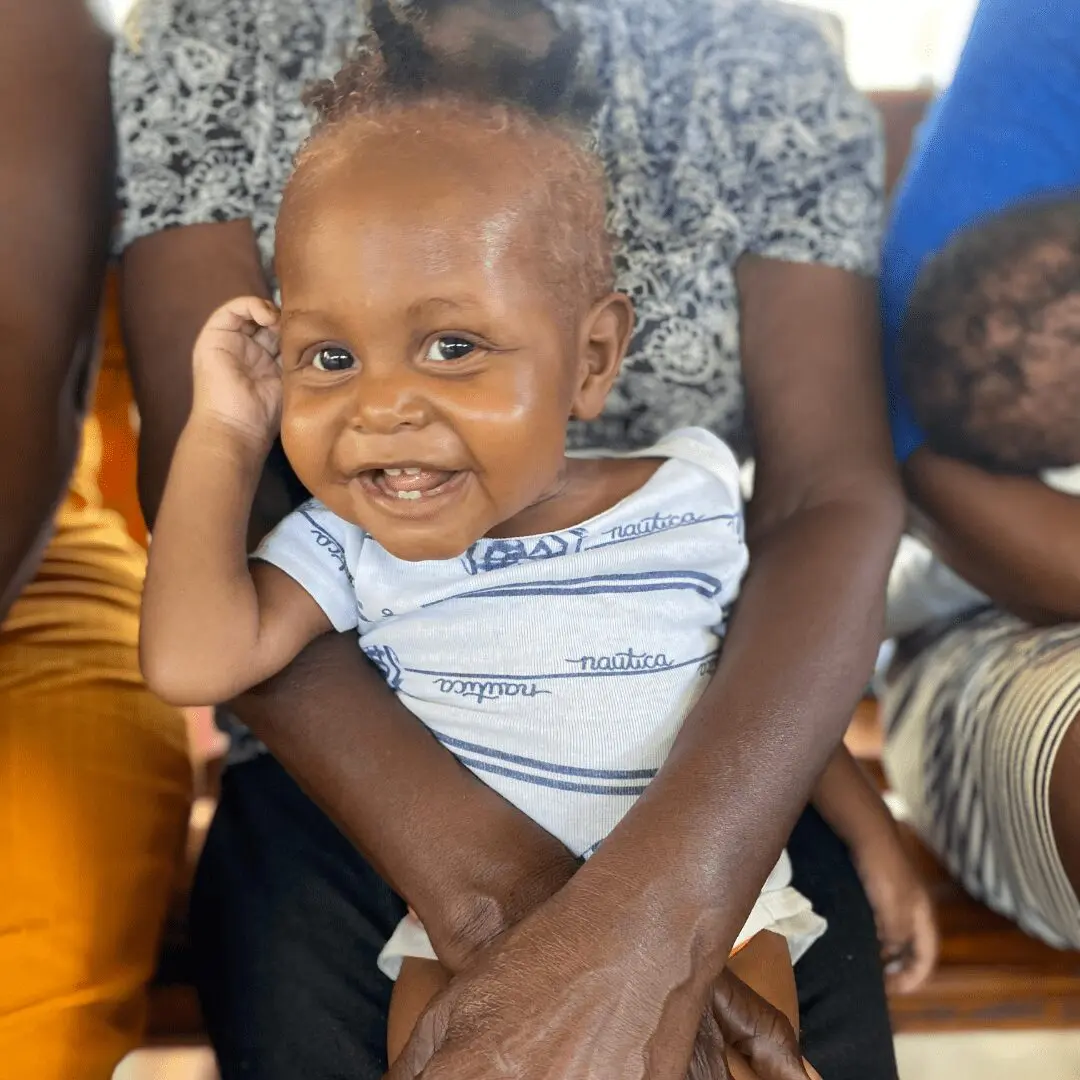 A close-up image shows a very happy Haitian infant in early recovery, with light reddish-brown hair, sitting on an adult's lap. The baby is wearing a white onesie with blue stripes and is looking directly at the camera with a wide, bright smile, one hand resting near his head. The adult's legs and shirt are visible, along with other people sitting nearby in the background.