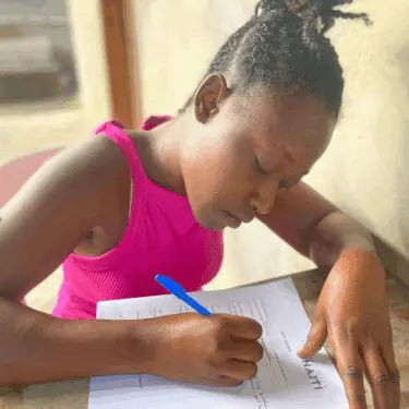 A young Haitian woman with her hair pulled back in twists concentrates intently as she writes on a document labeled "SECOND MILE HAITI" using a blue pen. She is wearing a bright pink tank top and leaning over a table in a brightly lit setting.
