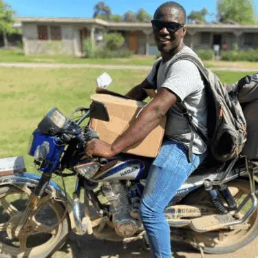 A smiling Haitian man wearing sunglasses, a gray t-shirt, and jeans sits on a blue and silver HAOJIN motorcycle, balancing a large cardboard box in front of him and wearing a backpack. He is outdoors in a sunny, possibly rural area, with houses and a grassy field in the background.