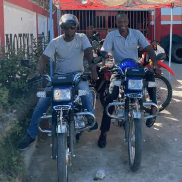 Two smiling Haitian men sit on new blue motorcycles, parked side-by-side in front of what appears to be a repair or sales shop. The man on the left wears a helmet and sunglasses, while the man on the right wears a polo shirt. Both bikes have 