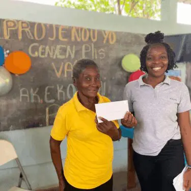 Two smiling Haitian women stand in front of a chalkboard with colorful writing in Haitian Creole and several balloons. The older woman on the left wears a bright yellow polo shirt and holds a white envelope, while the younger woman on the right wears a gray polo shirt with 