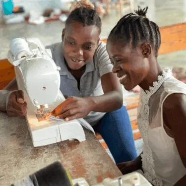 Two smiling Haitian women are engaged in a sewing lesson. The woman on the left, wearing a gray polo, leans forward to guide the fabric under the needle of a white sewing machine. The woman on the right, wearing a white sheer top with lace detailing, watches her with a bright smile. They are seated at a wooden table.