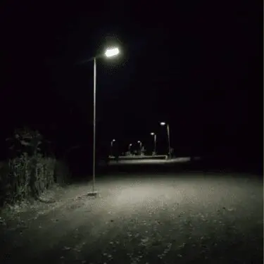 A dark image shows a solitary street lamp casting a bright white light onto a gravel or dirt path at night. The light illuminates the immediate foreground. Several other streetlights are visible in the distance along the path, receding into the darkness.