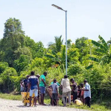 A group of men and boys stand around a yellow wheelbarrow on a dirt road, gathered near the base of a newly installed, tall solar-powered streetlamp. They are surrounded by lush, dense tropical vegetation under a bright, clear sky.